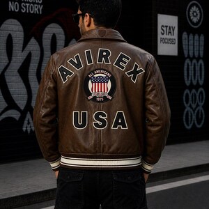 Puede incluir: Una chaqueta bomber de cuero marr&oacute;n con el texto AVIREX en arco en la parte superior de la espalda, un parche circular con una bandera estadounidense y el texto ALL AMERICANS ESTABLISHED 1975, y las letras USA en fuente blanca grande.