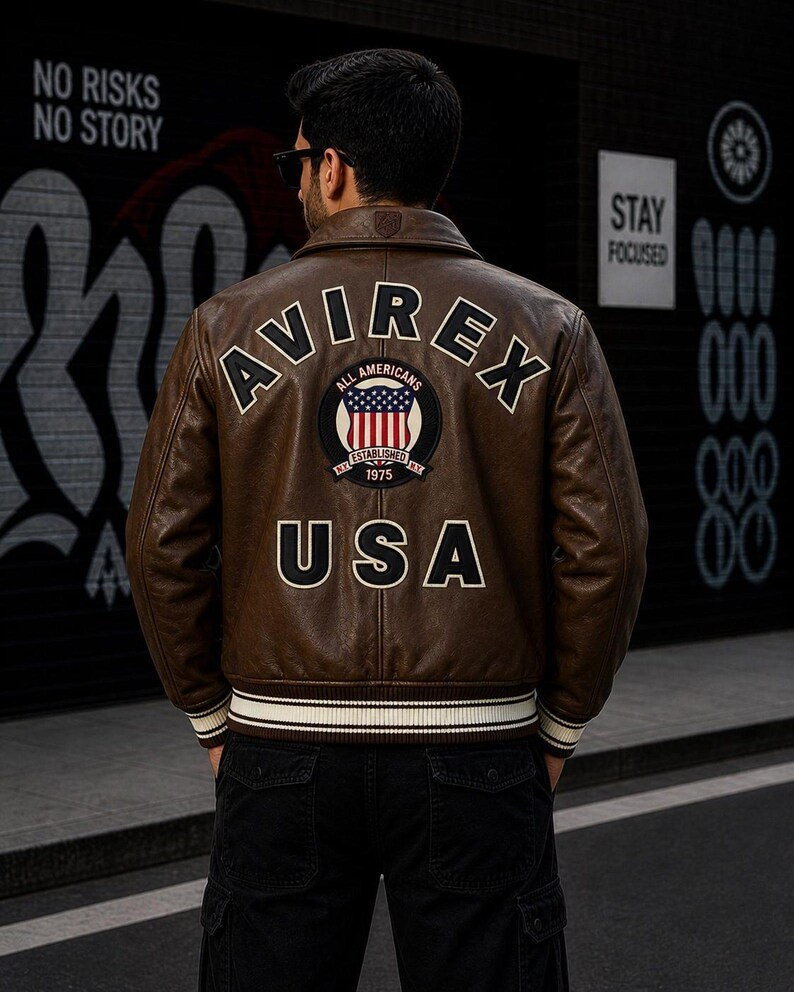 Puede incluir: Una chaqueta bomber de cuero marr&oacute;n con el texto AVIREX en arco en la parte superior de la espalda, un parche circular con una bandera estadounidense y el texto ALL AMERICANS ESTABLISHED 1975, y las letras USA en fuente blanca grande.