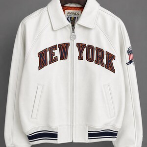 May include: A white varsity jacket featuring a full-length front zip, a fold-down collar, and ribbed cuffs and hem with navy blue stripes. The chest displays the text NEW YORK in navy blue and orange lettering, with an Avirex logo patch on the sleeve.