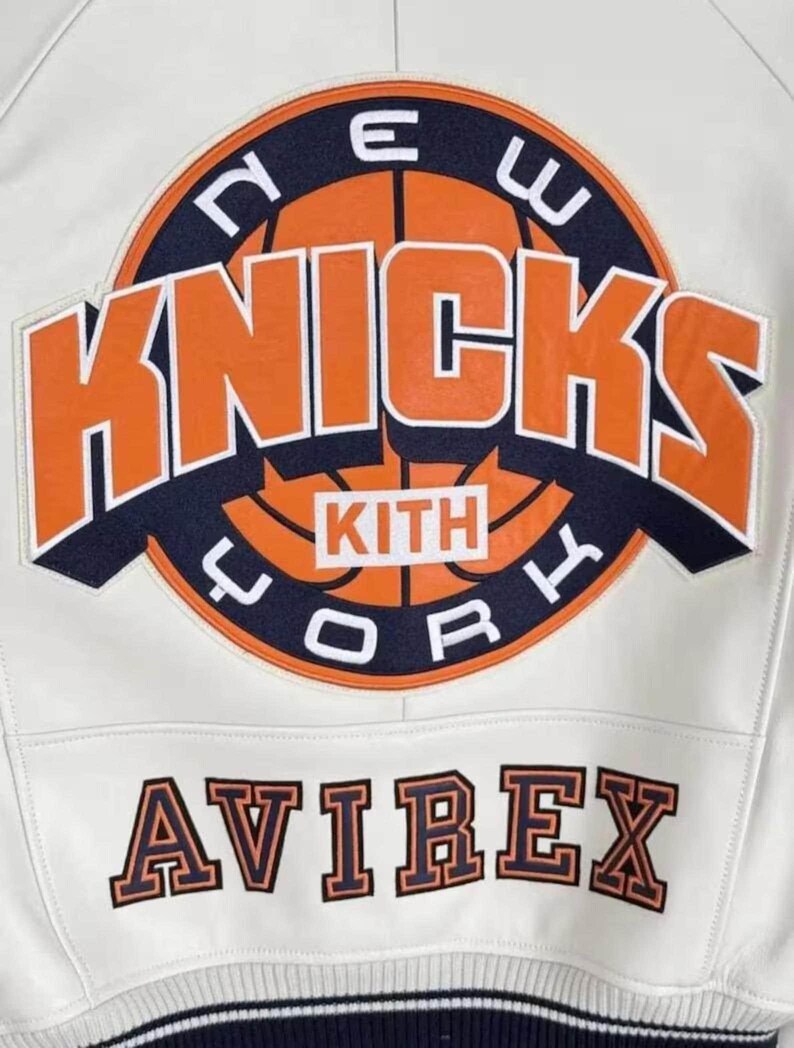 May include: A white leather jacket featuring a large circular embroidered patch on the back. The patch displays a basketball graphic with the text NEW YORK KNICKS and KITH in orange and navy blue. The word AVIREX is embroidered below in navy.