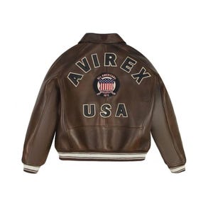 Puede incluir: Una chaqueta bomber de cuero marr&oacute;n con cuello, pu&ntilde;os y dobladillo acanalados con rayas blancas. La parte posterior muestra el texto AVIREX en arco, un parche circular con un dise&ntilde;o de bandera estadounidense y el texto ALL AMERICANS ESTABLISHED 1975, y USA.