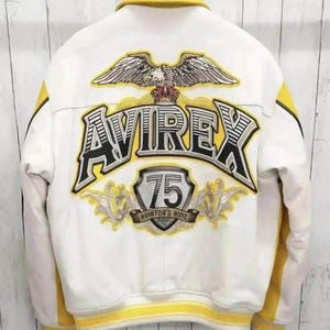 K&ouml;nnte beinhalten: Eine wei&szlig;-gelbe Collegejacke mit einer gro&szlig;en Stickerei auf der R&uuml;ckseite. Das Motiv zeigt einen Adler, den Schriftzug AVIREX, ein Wappen mit der Zahl 75 und den Text AVIATOR'S KING, alles in den Farben Schwarz, Wei&szlig; und Gelb gehalten.