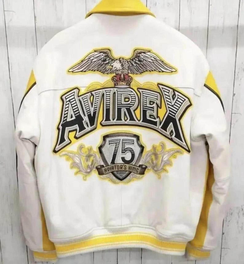 K&ouml;nnte beinhalten: Eine wei&szlig;-gelbe Collegejacke mit einer gro&szlig;en Stickerei auf der R&uuml;ckseite. Das Motiv zeigt einen Adler, den Schriftzug AVIREX, ein Wappen mit der Zahl 75 und den Text AVIATOR'S KING, alles in den Farben Schwarz, Wei&szlig; und Gelb gehalten.