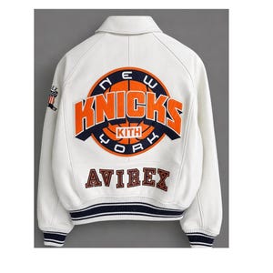 May include: A white varsity jacket featuring a large orange and navy blue New York Knicks basketball graphic on the back with the text KITH. The word AVIREX is embroidered below in navy blue. The cuffs and hem have navy blue and white stripes.