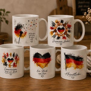May include: Five white ceramic mugs featuring watercolor-style designs with the German flag colors of black, red, and gold. The mugs display various illustrations including a dandelion, a heart, a flag, and people holding hands, with German text.