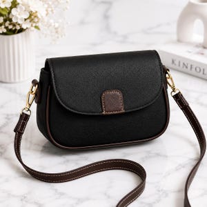 May include: A black crossbody bag with a textured finish, featuring a rounded flap closure with a dark brown accent tab. The bag includes gold-tone metal hardware, a dark brown adjustable shoulder strap with white stitching, and dark brown piping.