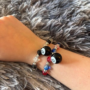May include: Two beaded bracelets featuring black billiard ball charms with the number eight printed in white. The bracelets are constructed with a mix of clear, black, red, and blue crackle glass beads interspersed with small metallic silver spacer beads.