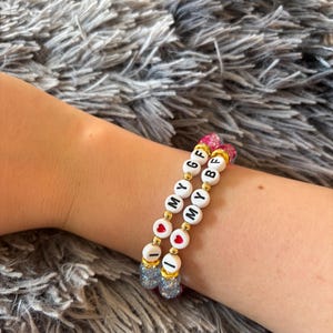 May include: Two beaded bracelets featuring white round beads with black letters and red heart symbols. One bracelet spells I MY GF and the other spells I MY BF. The design includes small gold-toned metallic spacer beads and translucent blue and pink textured beads at the ends.