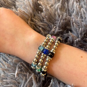 May include: Three beaded bracelets with silver-colored beads and colorful accent beads in blue, green, pink, and black. The bracelets are stacked on a wrist against a fuzzy gray background.