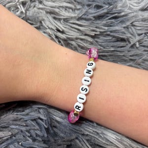 May include: A beaded bracelet featuring white round beads with the black printed letters R, O, S, I, N, G, interspersed with small gold-toned spacers and flanked by translucent, textured pink beads at both ends of the lettered sequence.