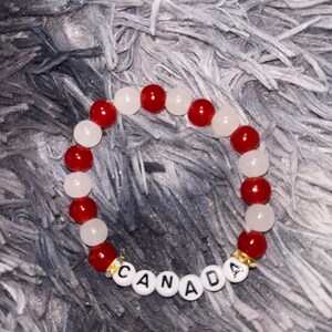 May include: A circular beaded bracelet featuring alternating red and white spherical beads. The bracelet includes white letter beads spelling out the word CANADA, flanked by two small gold-toned spacer beads with embedded clear crystals.