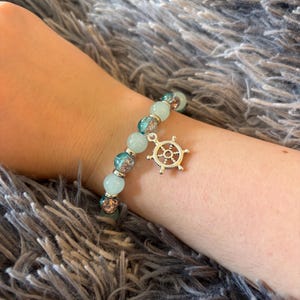 May include: A beaded bracelet featuring alternating smooth light blue beads and textured, glittery teal beads. The bracelet includes a silver-toned metal charm shaped like a ship's steering wheel, suspended from the strand of beads.
