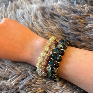 May include: A set of four stretch bracelets featuring round crackle glass beads in yellow, amber, teal, and black. Each bracelet is accented with small, metallic gold-toned spacer beads between the larger glass beads, creating a layered appearance.