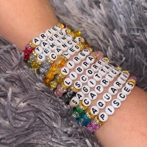 May include: A collection of beaded bracelets featuring zodiac signs. Each bracelet is made with white letter beads spelling out the zodiac names, interspersed with colorful beads and gold accents. The bracelets are stacked on a wrist.