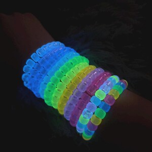 May include: A selection of vibrant, glowing bracelets in a rainbow of colours, including blue, green, yellow, and purple. The bracelets are made of small, round beads and are stacked on a wrist. The image is set against a dark background.
