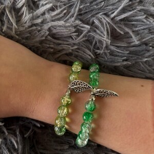May include: A double-strand bracelet featuring round crackle glass beads in shades of lime green and emerald green. The two strands are connected by a silver-toned metal charm shaped like a detailed leaf with intricate cut-out patterns.