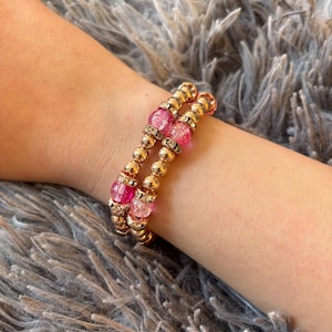 May include: A gold-colored beaded bracelet with pink and clear crystal accents. The bracelet is made of small, round gold beads and larger, faceted pink beads. The bracelet is worn on a wrist.