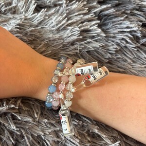 May include: Three beaded bracelets featuring round, crackled glass beads in shades of light blue, pale pink, and white. Each bracelet includes a miniature clear plastic bottle charm with a label that reads Bacardi Carta Blanca.