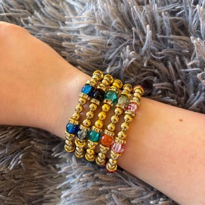 May include: Six gold-coloured beaded bracelets with colourful gemstone accents are stacked on a wrist. The gemstones are blue, black, teal, green, orange, and pink. The bracelets have a mix of small gold beads and larger gemstone beads.