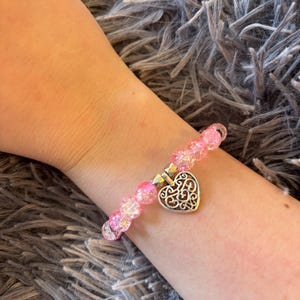 May include: A pink beaded bracelet with a silver heart charm. The bracelet features clear, iridescent beads and a decorative heart pendant. The heart charm has an openwork design.
