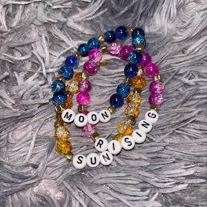 May include: Two elastic beaded bracelets featuring crackle-textured glass beads in shades of blue, pink, and yellow, accented with small gold-toned spacers. White letter beads spell out the words MOON RISING and SUN RISING on the bracelets.