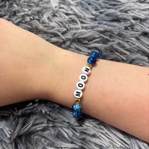 May include: A beaded bracelet featuring round, dark blue, textured beads alternating with small gold-toned square spacers. The centre of the bracelet displays four white, round, flat beads with black letters spelling out the word MOON.