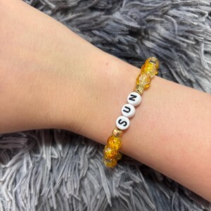 May include: A beaded bracelet featuring yellow crackle glass beads, small gold-toned metallic spacers, and three white circular beads with the black printed letters S, U, and N, arranged to spell the word SUN in the centre of the elastic band.