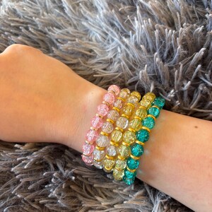 May include: Five beaded bracelets in pink, yellow, and teal are displayed on a wrist. The round, faceted beads are separated by gold-coloured spacers. The bracelets are worn against a grey, textured background.