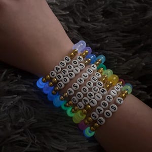 May include: A stack of nine elastic bracelets featuring colourful translucent beads and gold-toned metallic spacers. Each bracelet includes white letter beads spelling out the names Cass, Brook, Doris, Ashley, Cooper, Lance, Tim, Sabrina, and Rosa.