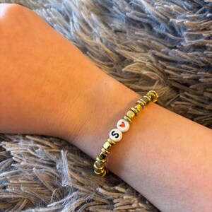 May include: A stretch bracelet featuring metallic gold-toned disc beads interspersed with two white circular beads. One white bead displays the black letter S, while the other white bead features a small red heart symbol centred on the surface.