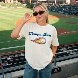 Könnte beinhalten: Ein cremefarbenes Kurzarm-T-Shirt mit dem Schriftzug Tampa Bay in blauer Schreibschrift über einer Grafik eines Baseballs, der von orangefarbenen, gelben und roten Flammen umgeben ist, kombiniert mit einer blauen Jeans und einer dunklen Sonnenbrille.