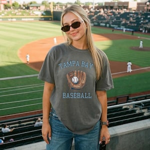May include: A charcoal grey short-sleeve t-shirt featuring a graphic of a baseball glove holding a baseball. The text TAMPA BAY is printed in light blue above the graphic, and the word BASEBALL is printed in light blue below the graphic.