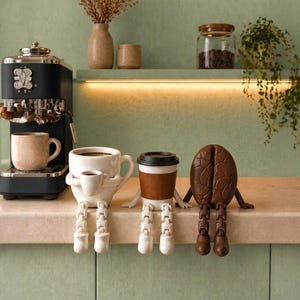 Peut inclure: Trois figurines décoratives assises sur un rebord : une tasse blanche avec une plus petite tasse attachée, un gobelet blanc avec un manchon marron et un couvercle noir, et une forme de grain de café marron. Chaque figurine a des jambes articulées.