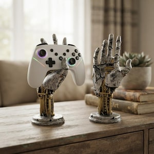 Op de afbeelding: Twee mechanische handsculpturen met een verweerde zilveren en gouden afwerking, met zichtbare interne tandwielen en bedrading. De ene hand staat rechtop, terwijl de andere een witte draadloze gamecontroller vasthoudt.