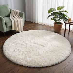 May include: A large, circular, off-white area rug with a thick, shaggy, faux-fur texture. The rug is positioned on a dark brown wooden floor, centered in the room, featuring a soft, plush pile that covers the floor surface in a round shape.