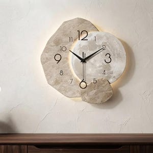 Peut inclure: Une horloge murale au design en pierre irrégulière et superposée dans des tons beige et crème. Le cadran affiche des chiffres noirs et des aiguilles fines, avec un pendule circulaire en bas et un rétroéclairage intégré.