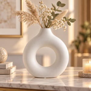 Puede incluir: Un jarrón de cerámica blanco mate con un cuerpo circular en forma de rosquilla y una abertura central. El jarrón contiene un arreglo floral seco compuesto por hierba de la pampa beige, pequeñas flores blancas de paniculata y hojas de eucalipto verdes.
