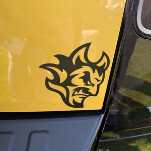 May include: A black vinyl decal featuring a stylised devil head with flame-like hair and horns, applied to the yellow painted surface of a vehicle. The graphic is positioned near the black trim and rear light assembly of the car body panel.