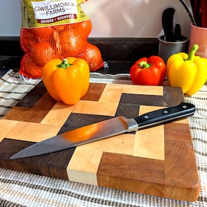 May include: A rectangular wooden chopping board featuring a geometric herringbone pattern made of alternating light and dark wood blocks. A stainless steel chef knife with a black handle rests diagonally across the surface of the board.