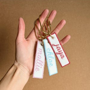 May include: Three rectangular white fabric keychains attached to a gold-toned metal ring. Each keychain features embroidered names in cursive script: one in pink thread reading mollie, one in light blue reading chloe, and one in red reading jaya.