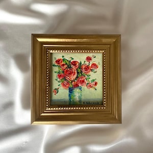 Könnte beinhalten: Ein quadratisches Ölgemälde in einem goldenen Rahmen zeigt einen Strauß roter Rosen in einer blauen Vase vor hellgrünem Hintergrund. Das Kunstwerk weist dicke, strukturierte Pinselstriche auf und trägt unten rechts die Signatur A. Valenzuela.