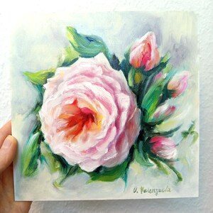 Könnte beinhalten: Ein Ölgemälde auf quadratischer Leinwand, das eine große, blühende blassrosa Rose mit einem tiefroten und gelben Zentrum zeigt, umgeben von mehreren kleineren rosa Rosenknospen und grünen Blättern. Die Signatur V. Valenzuela steht unten rechts.