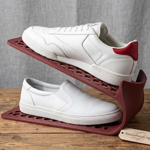 Op de afbeelding: Een donkerrood plastic schoenenrek met een gelaagd, open ontwerp met een honingraatpatroon. Het rek houdt een witte sneaker met veters en een rood hielaccent op de bovenste laag en een witte instapschoen op de onderste laag.