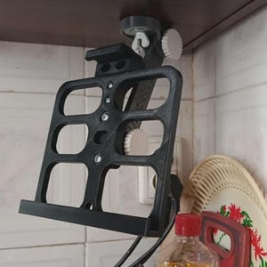 May include: A black, adjustable, wall-mounted cookbook holder. The holder is made of a dark material and features a rectangular frame with multiple openings. It is attached to a shelf with a clamp and adjustable knobs. A bottle and a decorative plate are visible below.