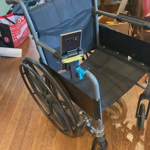 May include: A grey metal wheelchair with a black fabric seat and backrest, featuring a black smartphone mounted to the left armrest using a black plastic holder secured with a yellow rubber band and a small blue clamp component.