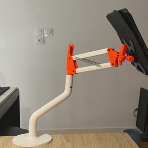 Puede incluir: Un soporte ajustable para tableta en blanco y naranja con base circular, brazo de soporte curvo y mecanismo de extensión multiarticulado. El dispositivo cuenta con un soporte de montaje que sujeta una tableta negra inclinada.