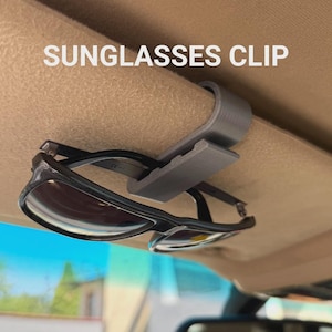 Op de afbeelding: Een grijze plastic zonnebrilclip bevestigd aan een beige zonneklep in de auto, met daarin een zonnebril met een zwart montuur. De tekst SUNGLASSES CLIP staat in het wit erboven gedrukt. Het apparaat heeft een U-vormig ontwerp voor bevestiging.