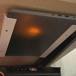 Może przedstawiać: Ciemnoszary laptop zamontowany poziomo w jasnym uchwycie do przechowywania pod biurkiem. Uchwyt posiada dwie równoległe szyny boczne z widocznymi otworami na śruby, które pewnie mocują urządzenie pod płaską powierzchnią blatu biurka.
