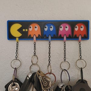 Könnte beinhalten: Ein rechteckiger Wand-Schlüsselhalter mit schwarzer Platte und blauem Rand. Er zeigt eine gelbe Pac-Man-Figur und vier bunte Geister in Orange, Blau, Pink und Rot. Jede Figur hat eine hängende schwarze Kette mit einem metallenen Schlüsselring.