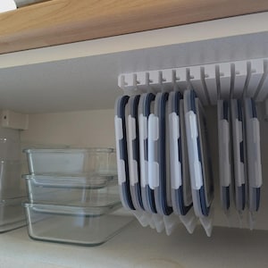 May include: A white plastic lid organiser with multiple hanging lids in a kitchen cupboard. The lids are blue and white, and there are several clear glass food storage containers on the shelf below.